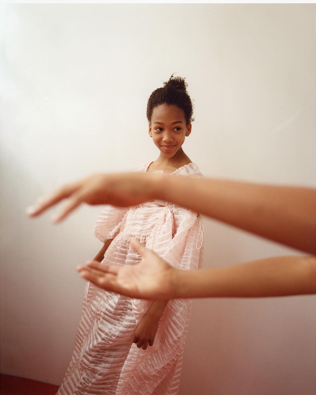 Young girl in a pink dress with sheer, ruffled fabric stands near a person's extended hands, creating a dynamic and engaging portrait.