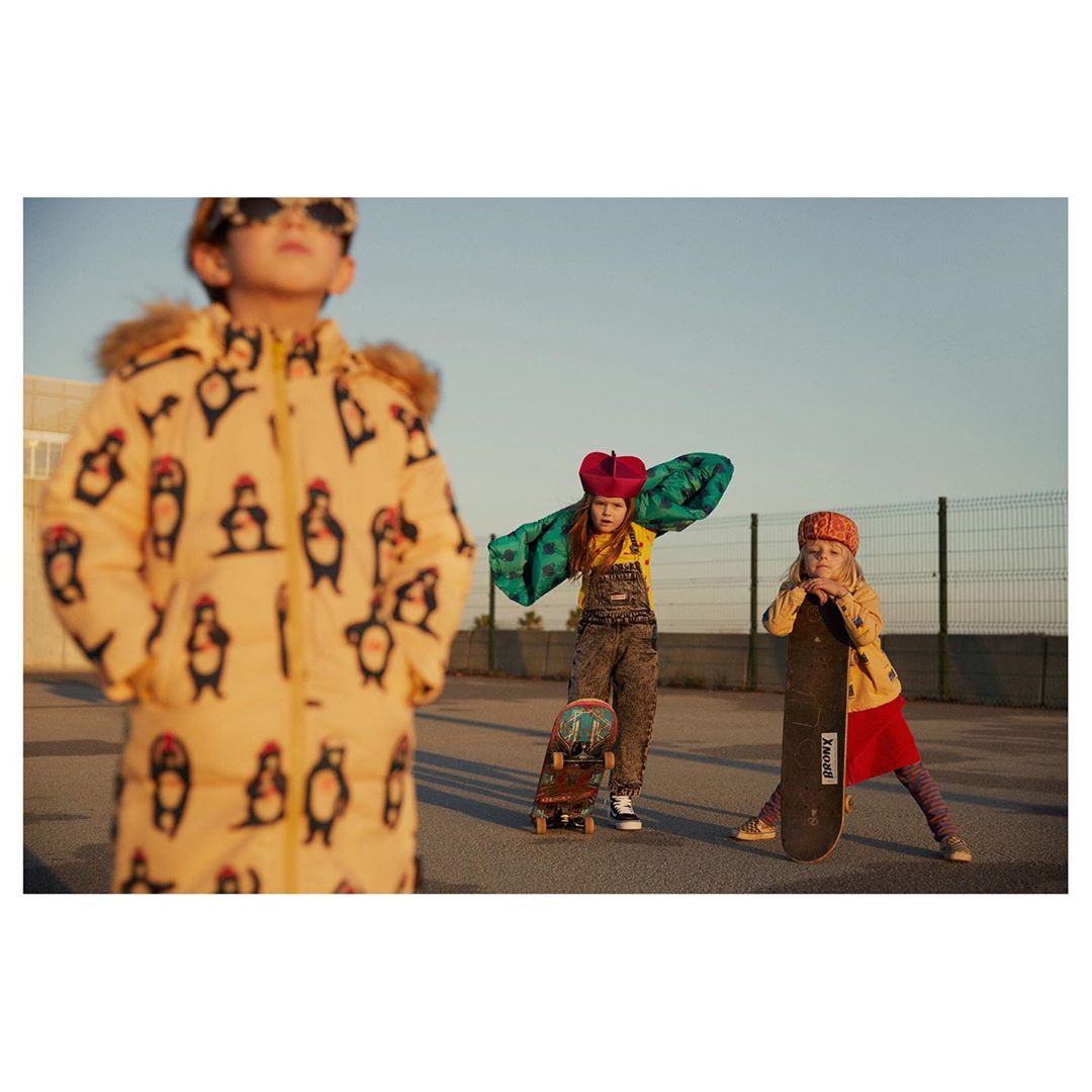 Three children stand outdoors on a sunny day; one wears a penguin-patterned coat, another is in overalls with a green shawl, with skateboards.