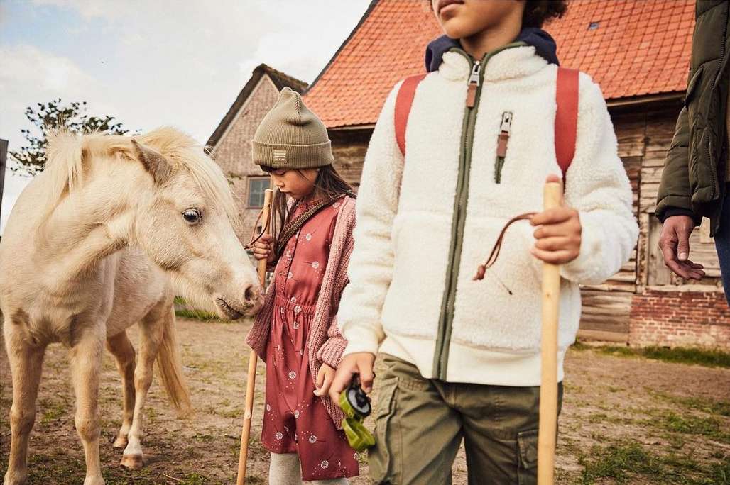 Children and a pony stand near a rural barn, dressed in outdoor clothing with earthy tones, evoking a lifestyle fashion editorial.