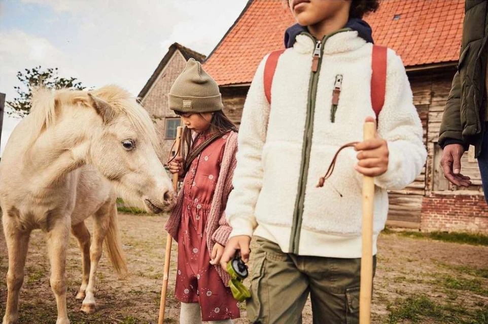 Children and a pony stand near a rural barn, dressed in outdoor clothing with earthy tones, evoking a lifestyle fashion editorial.