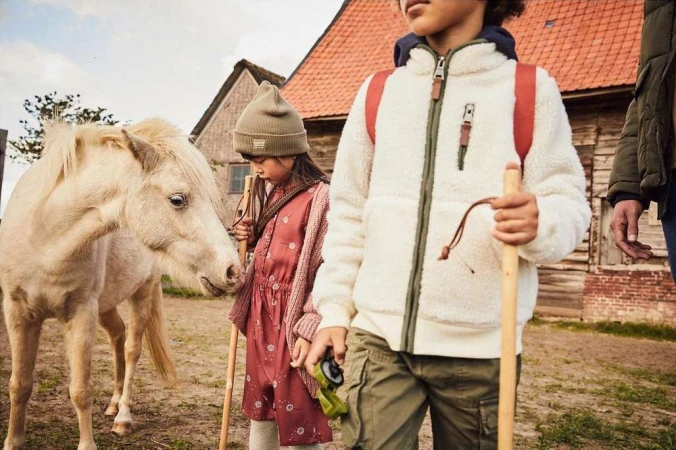 Children and a pony stand near a rural barn, dressed in outdoor clothing with earthy tones, evoking a lifestyle fashion editorial.