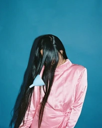 Woman in pink satin dress with long black hair obscuring her face and a blue funnel tucked into her hair against a blue background