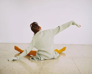 A person with braided hair wearing a sage green top and yellow tights stretches on a tiled floor against a white wall.