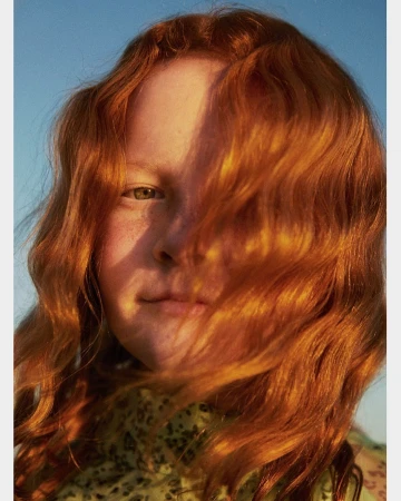 Close-up portrait of a woman with vibrant red hair partially covering her face against a clear blue sky background