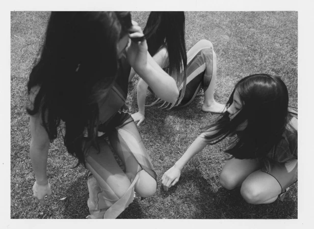 Three young women with long dark hair kneeling on a grassy lawn outdoors photographed from a high angle in black and white.