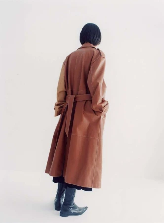 Model with short dark hair viewed from behind wearing a long brown belted coat and dark boots against a white studio background.
