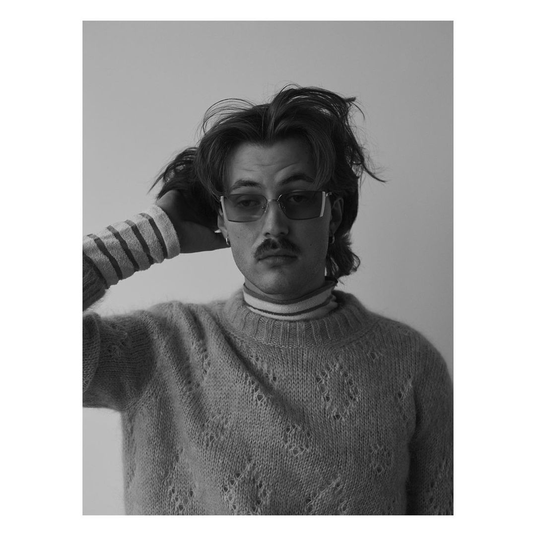 Black and white portrait of a stylish man with a mustache, wearing sunglasses and a patterned sweater, posing in a photo studio.