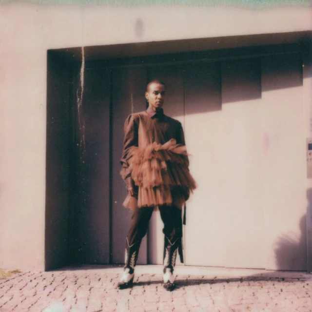 A person wearing a tulle garment and cowboy boots stands in front of a gray door in a medium shot, outdoors on a brick surface.