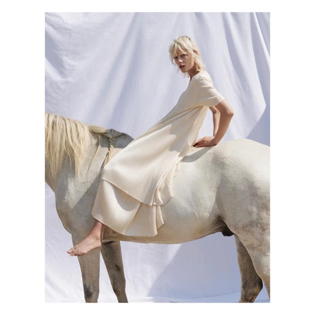 Adult female model in a long pleated dress is posing bareback on a white horse in an indoor photo studio with a white backdrop.