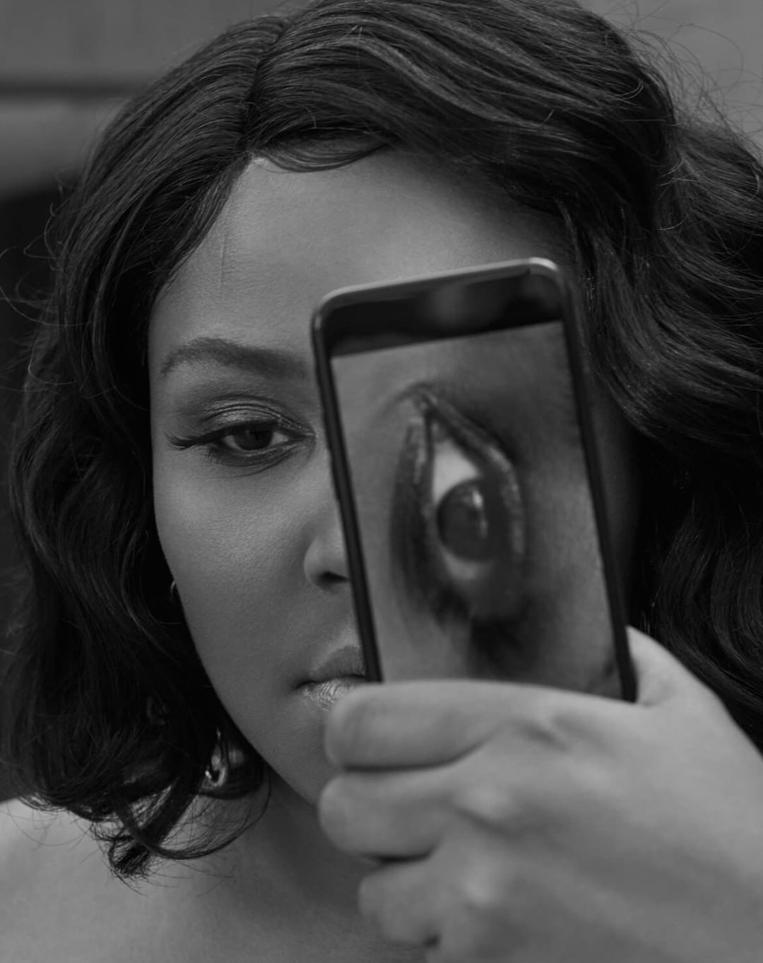 Black and white portrait of a woman holding a smartphone up to her face, displaying a close-up image of an eye on screen.