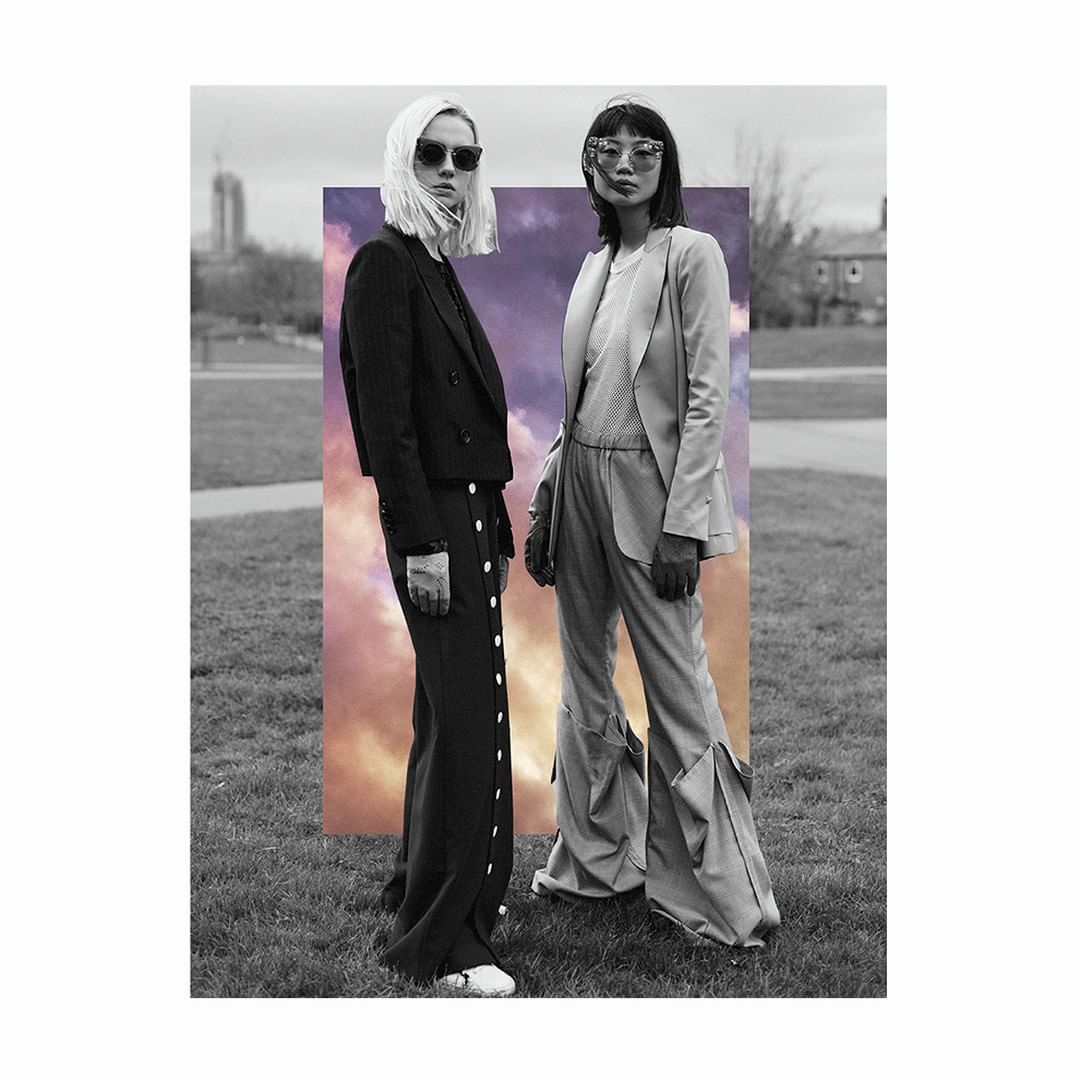 Two fashion models in matching suits pose in a field with a cloud backdrop and a black-and-white urban park.  