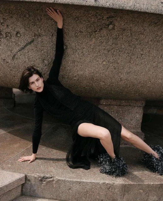 Fashionable woman in a black dress with a side slit poses on stone steps with a textured stone background, showcasing a unique style.