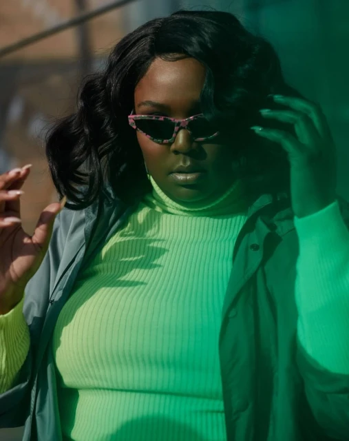 A stylish woman with sunglasses in a neon top poses in dappled light, showcasing a fashion-forward look with dark hair and a jacket.
