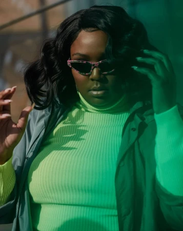 A stylish woman with sunglasses in a neon top poses in dappled light, showcasing a fashion-forward look with dark hair and a jacket.