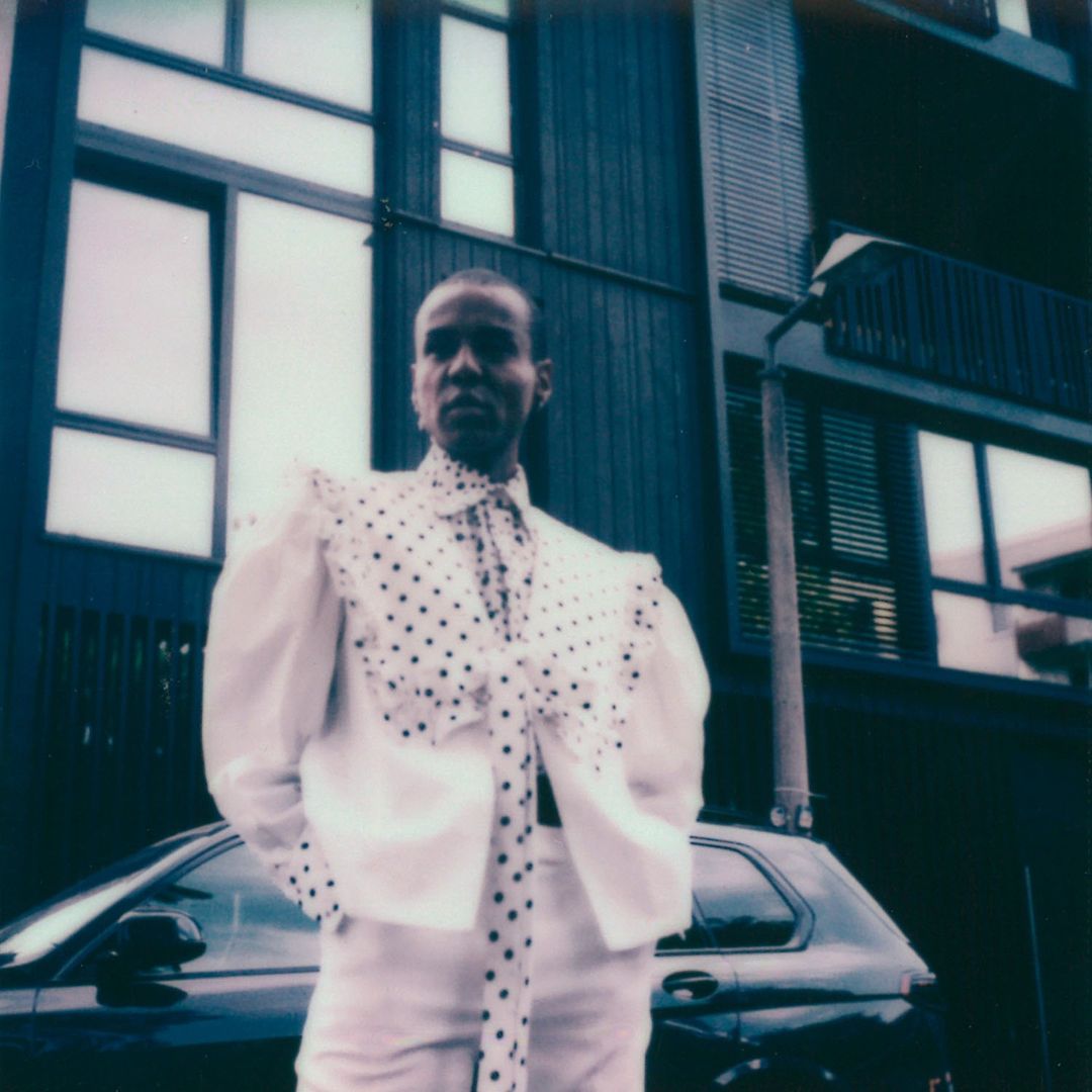 Adult female in white polka-dot jacket and pants poses by black car near modern building in urban environment, fashion style, daytime.