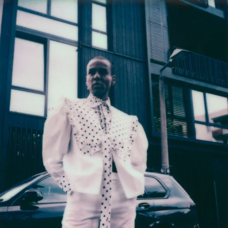 Adult female in white polka-dot jacket and pants poses by black car near modern building in urban environment, fashion style, daytime.