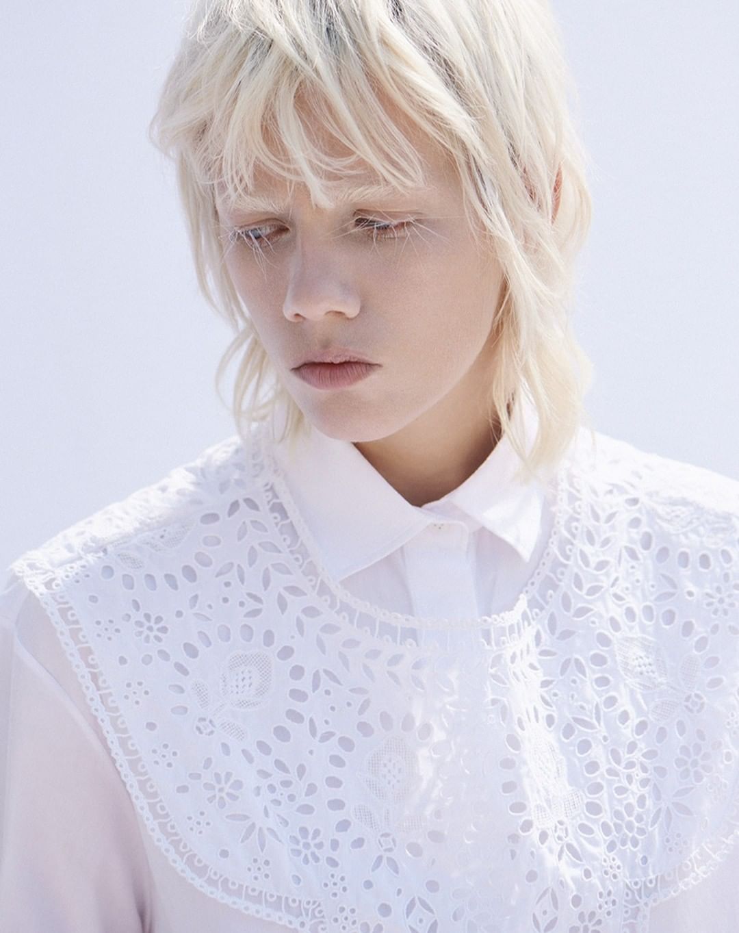 Young woman with platinum blonde hair, wearing an openwork collar, poses indoors against a bright background in a portrait shot.