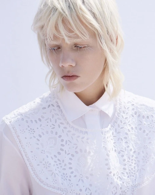 Young woman with platinum blonde hair, wearing an openwork collar, poses indoors against a bright background in a portrait shot.