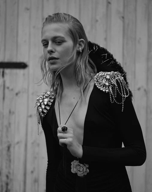Monochrome portrait of a blonde woman wearing an embellished black dress, holding a ring, against a wooden wall backdrop.