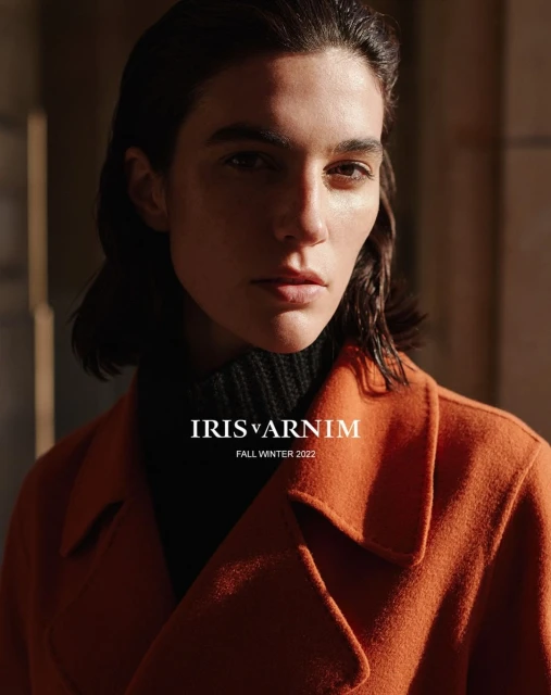 A fashion shot features a brunette adult female in an orange coat, set against a textured background, illuminated by warm lighting.