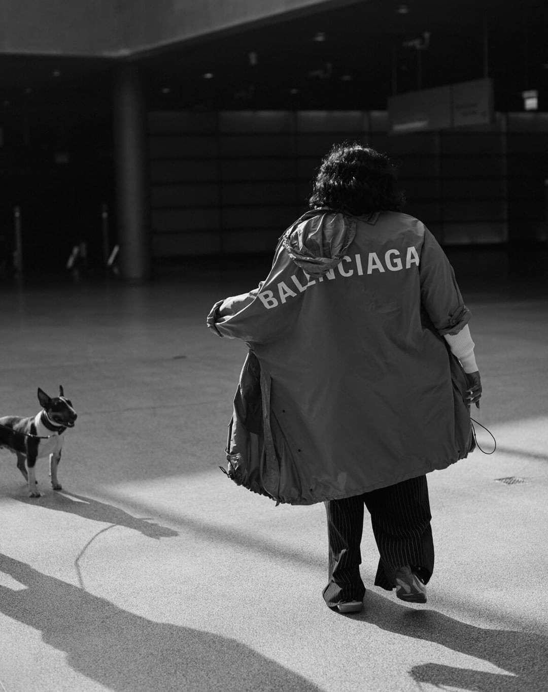 Person wearing a Balenciaga branded jacket walking a dog on a leash across a polished concrete floor.