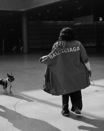 Person wearing a Balenciaga branded jacket walking a dog on a leash across a polished concrete floor.