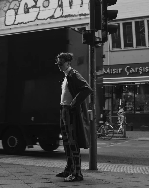 Monochrome portrait of a slender adult male standing on a city sidewalk with a traffic light, truck, and building in the background.