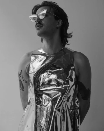 Man wearing angular geometric sunglasses and a reflective metallic top against a plain light background