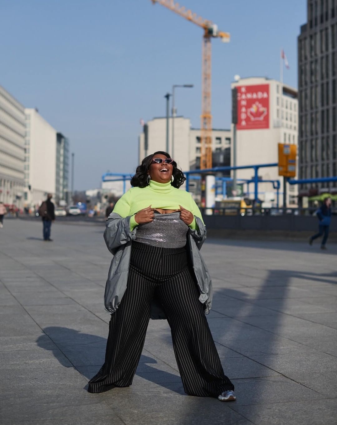 Adult woman poses outdoors in a neon turtleneck, silver sequin top, and black striped pants in an urban area on a sunny day.