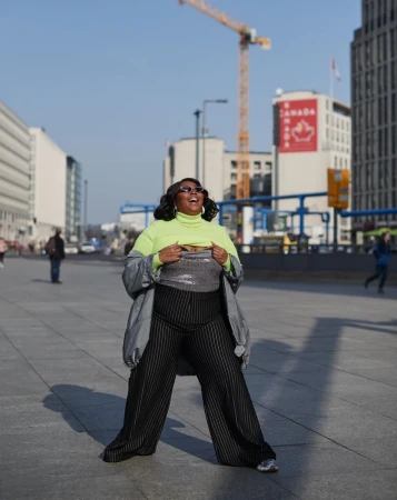 Adult woman poses outdoors in a neon turtleneck, silver sequin top, and black striped pants in an urban area on a sunny day.