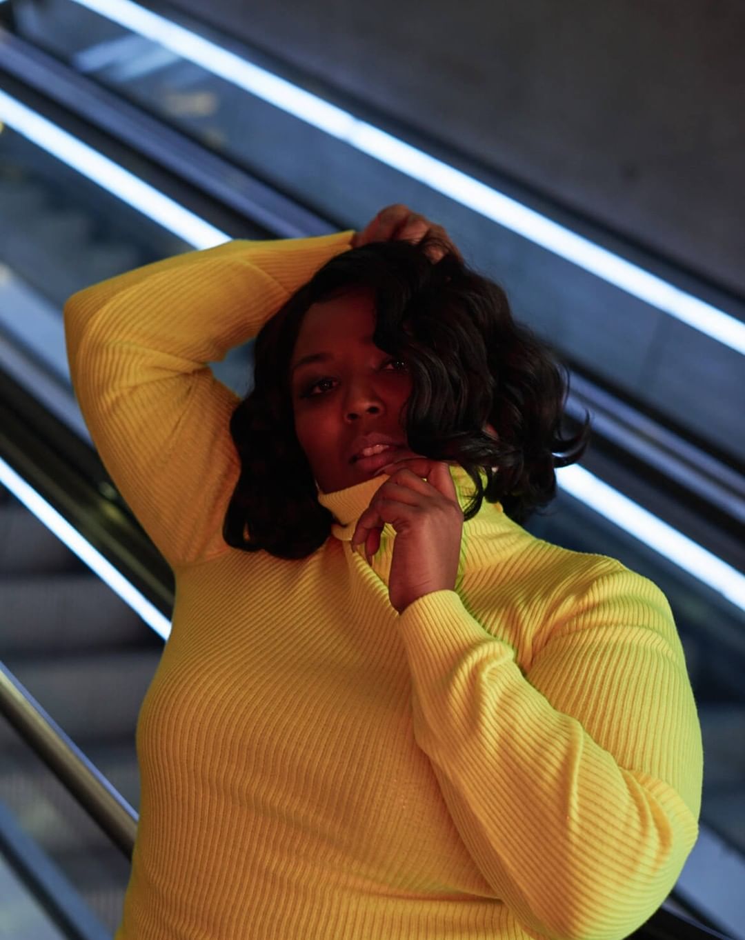 Portrait of a Black woman in a yellow turtleneck sweater posing in front of a background of illuminated diagonal lines indoors.