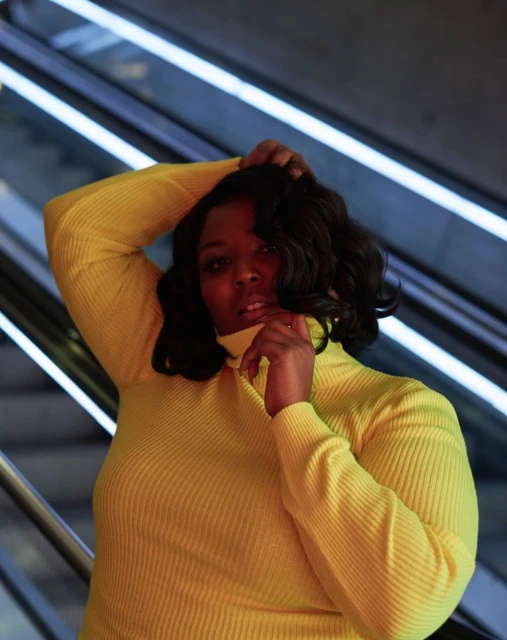 Portrait of a Black woman in a yellow turtleneck sweater posing in front of a background of illuminated diagonal lines indoors.