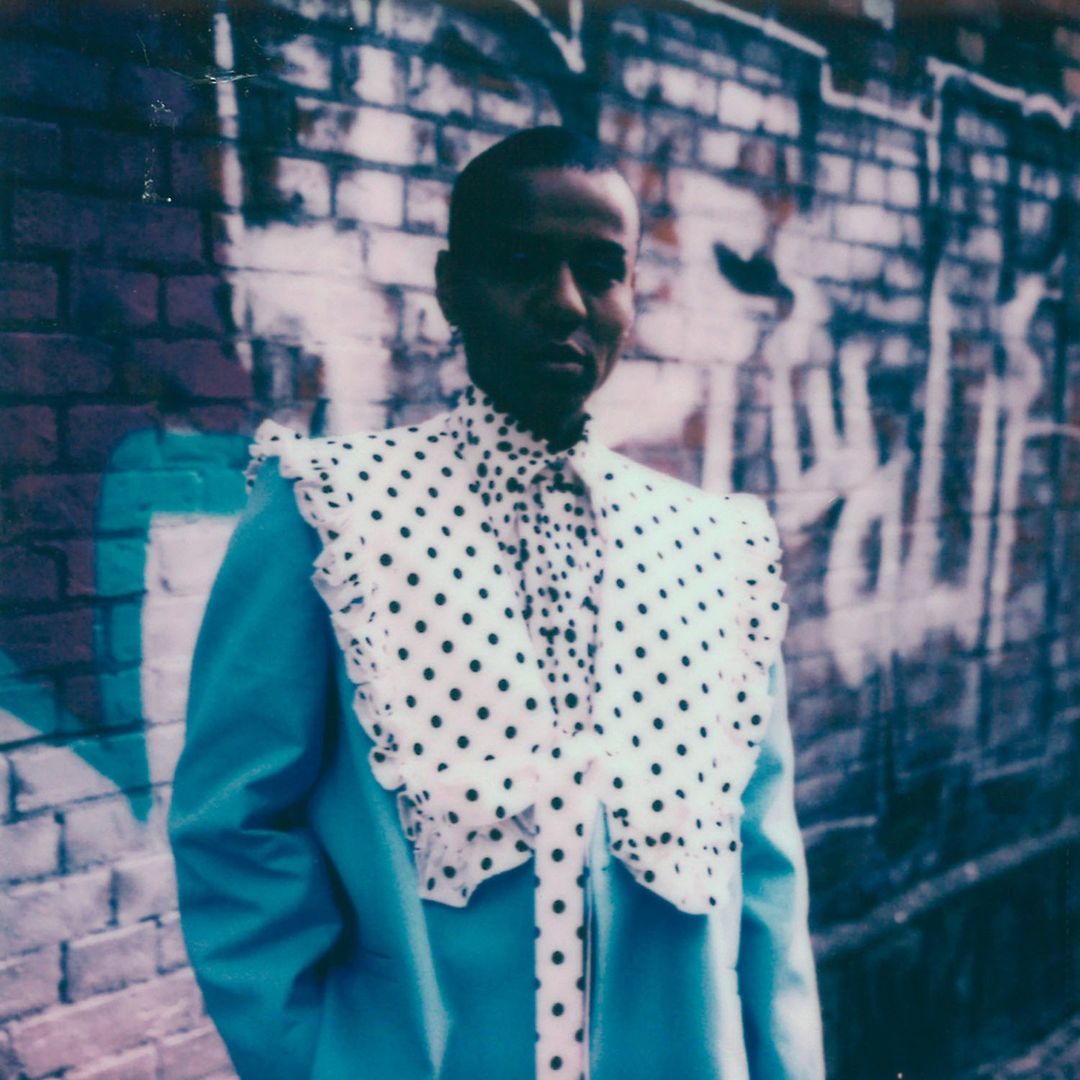 Person in a turquoise coat with a white polka dot ruffle collar and shirt, poses in front of an old red brick wall with graffiti.