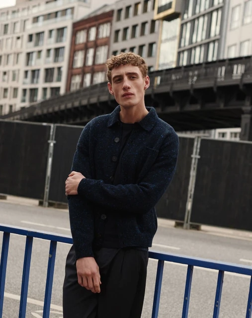An adult male with curly brown hair stands on a city street wearing a navy blue jacket and black pants, posing in front of buildings.
