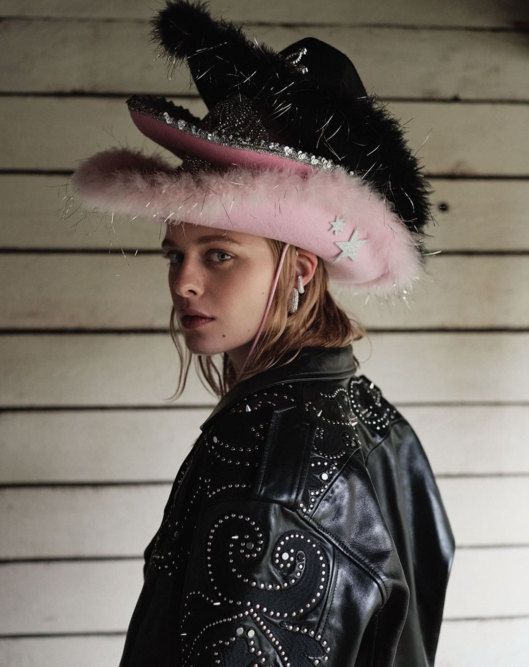 Portrait of a female model wearing a black studded leather jacket and a colorful, layered cowboy hat in front of a weathered wall.