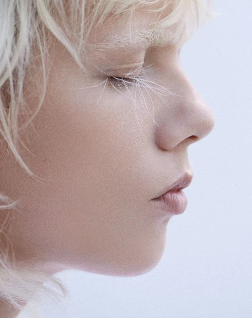 Close-up profile portrait of a fair-skinned person with white-blonde hair and decorative white feather eyelashes against a soft, light-blue background.