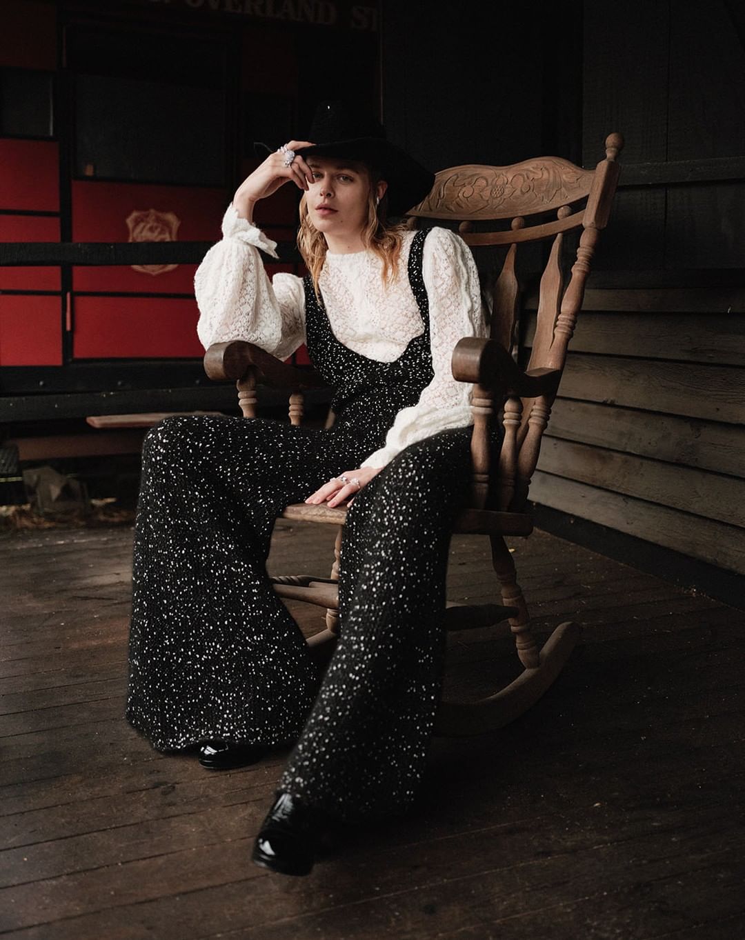 Woman in a black hat and jumpsuit sitting in a wooden rocking chair indoors on a wooden floor, posing for a fashion shot.