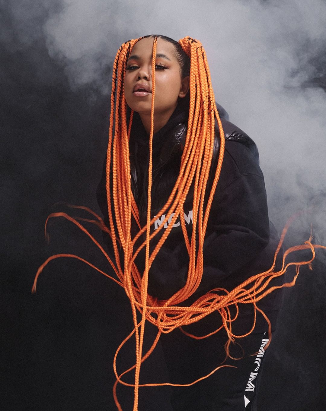 Stylish young woman with orange braided pigtails poses in a black hoodie against a smoky gray backdrop in a studio shot.