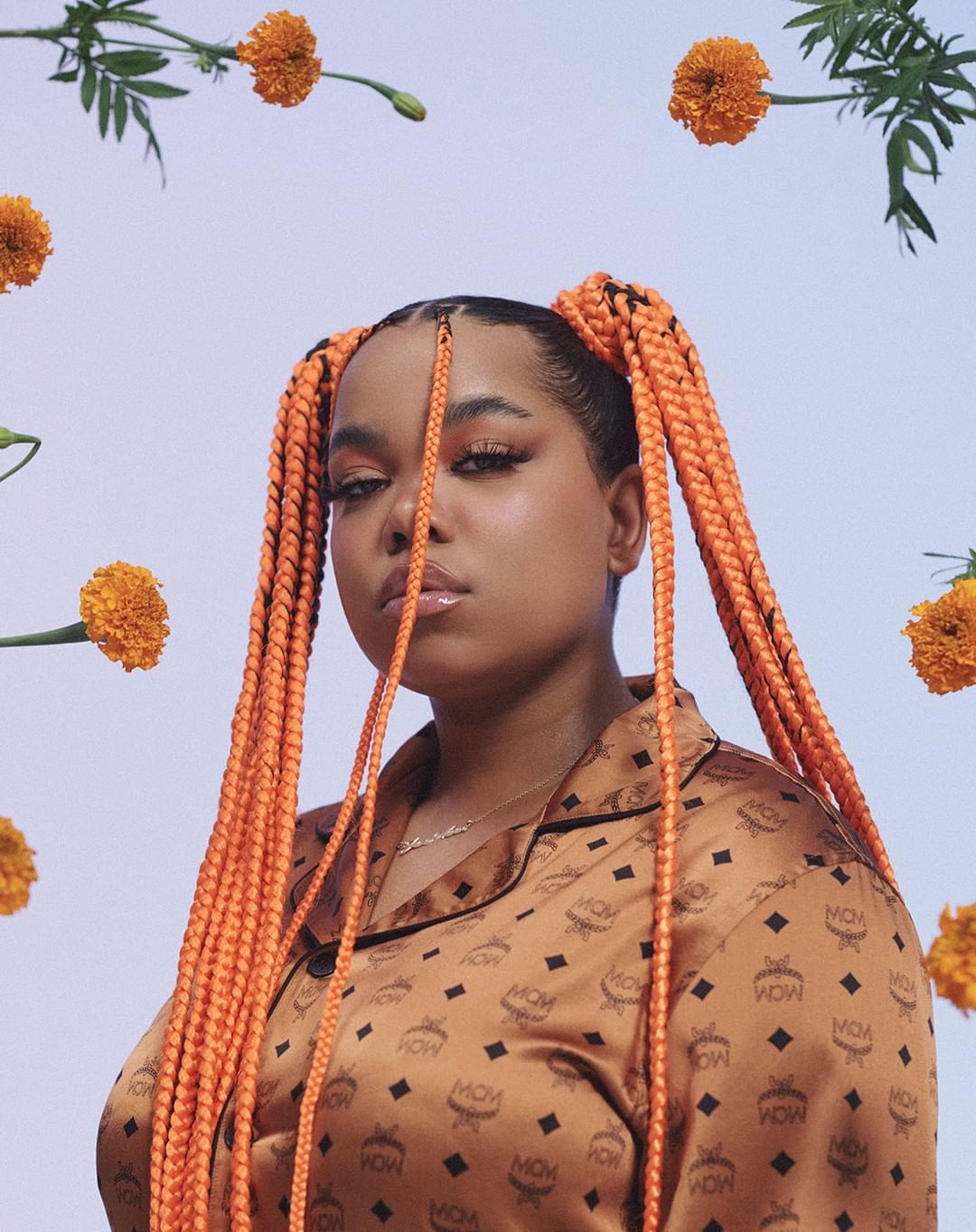 Portrait of a woman with braided pigtails, wearing a patterned blouse, framed by orange flowers against a light violet background.