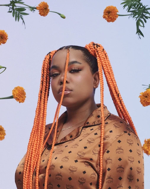 Portrait of a woman with braided pigtails, wearing a patterned blouse, framed by orange flowers against a light violet background.