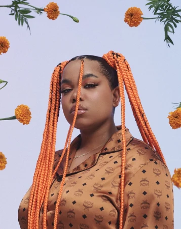 Portrait of a woman with braided pigtails, wearing a patterned blouse, framed by orange flowers against a light violet background.