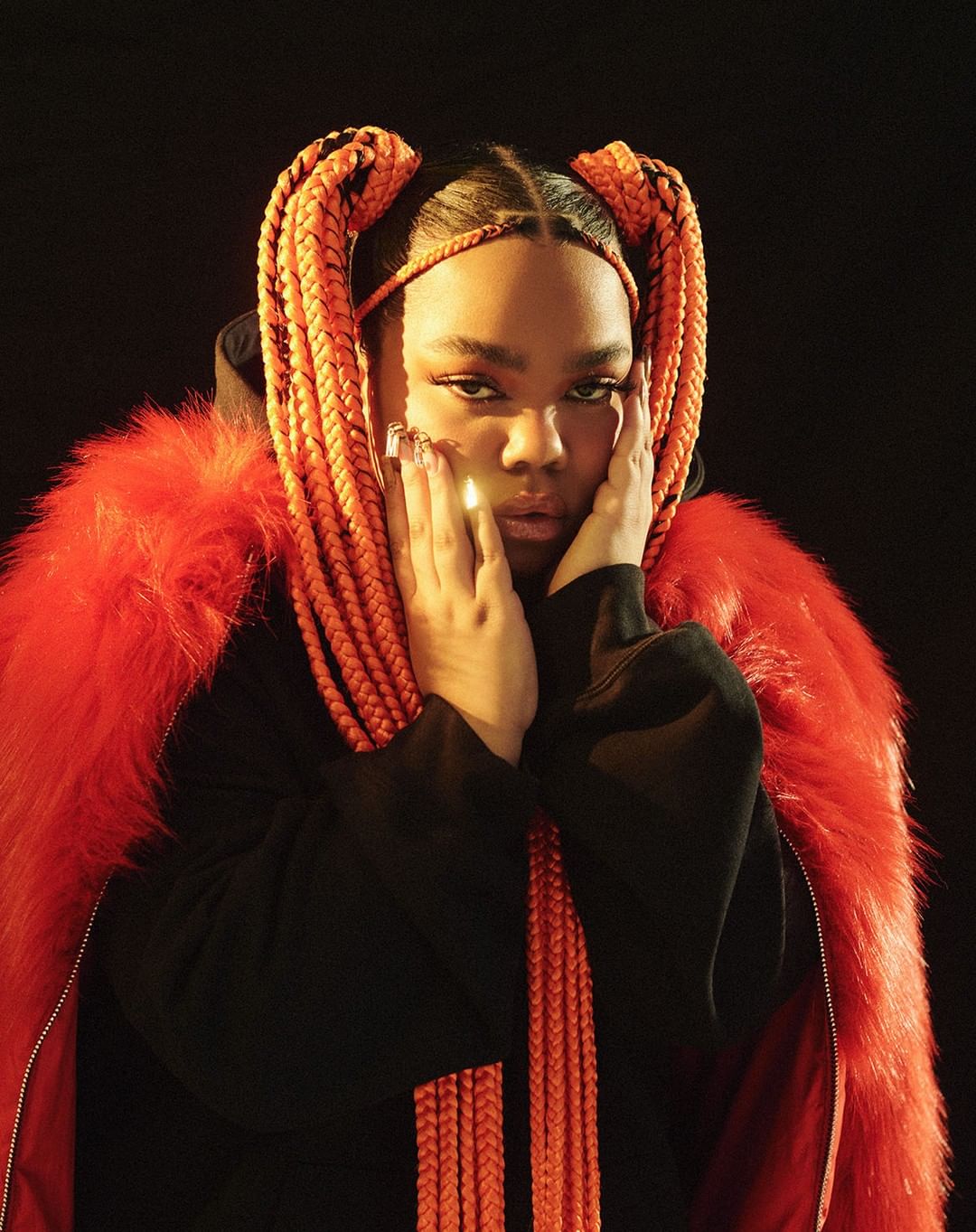 Portrait of a woman with orange braided pigtails and a red fur-trimmed jacket, looking at the camera against a dark background.
