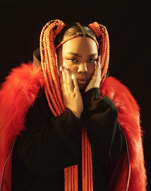 Portrait of a woman with orange braided pigtails and a red fur-trimmed jacket, looking at the camera against a dark background.