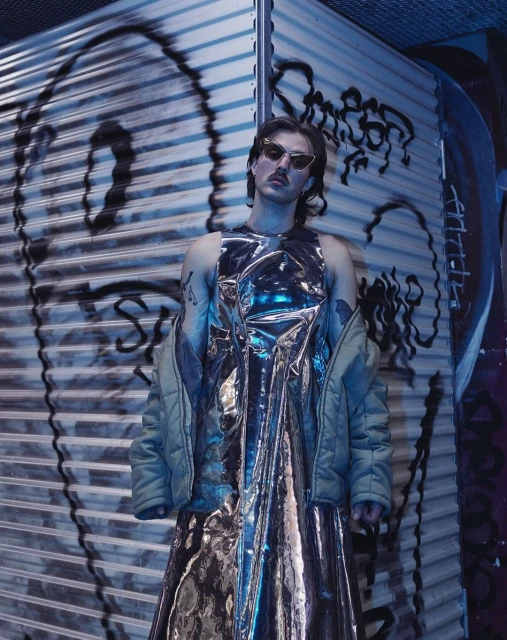 Person in metallic dress poses in front of graffiti on corrugated metal wall wearing sunglasses and holding a jacket. 
