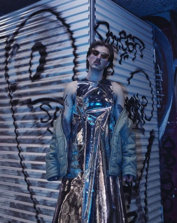 Person in metallic dress poses in front of graffiti on corrugated metal wall wearing sunglasses and holding a jacket. 