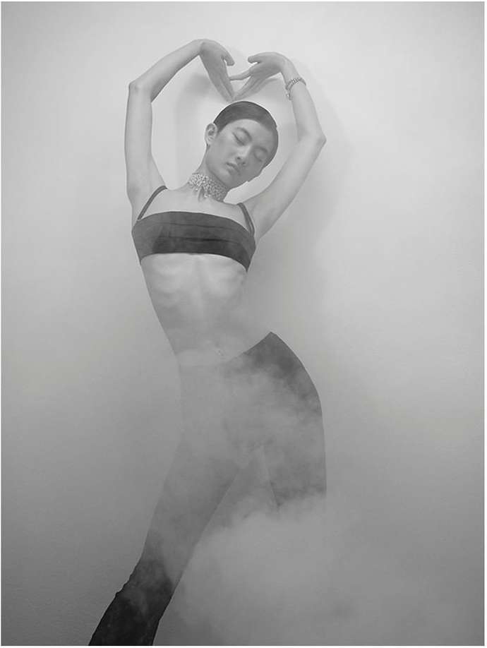 Monochrome portrait of a person posing in a dark bandeau top, bracelet, and choker. The person is surrounded by haze and a light background.