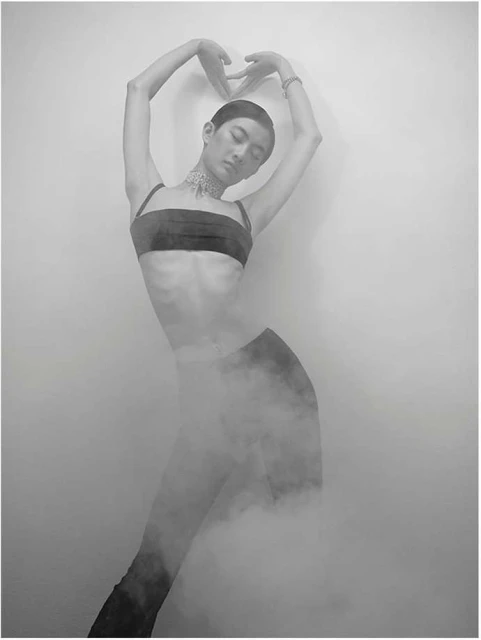 Monochrome portrait of a person posing in a dark bandeau top, bracelet, and choker. The person is surrounded by haze and a light background.