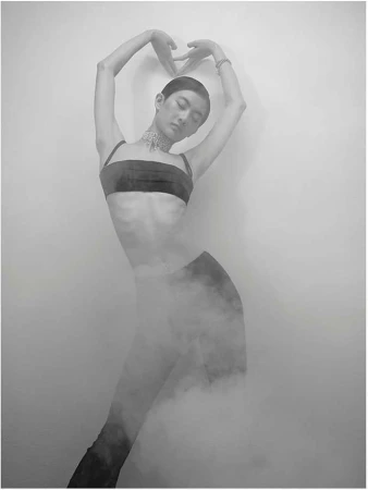Monochrome portrait of a person posing in a dark bandeau top, bracelet, and choker. The person is surrounded by haze and a light background.