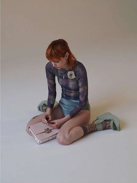 Young woman with auburn hair sits on the floor next to a light pink purse and wears a long-sleeved top and teal-colored leotard.