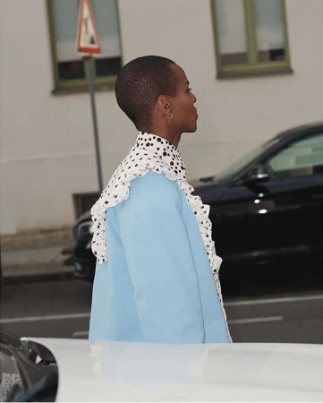 Profile of a person with short hair wearing a light blue jacket and polka dot ruffled collar standing on a street.